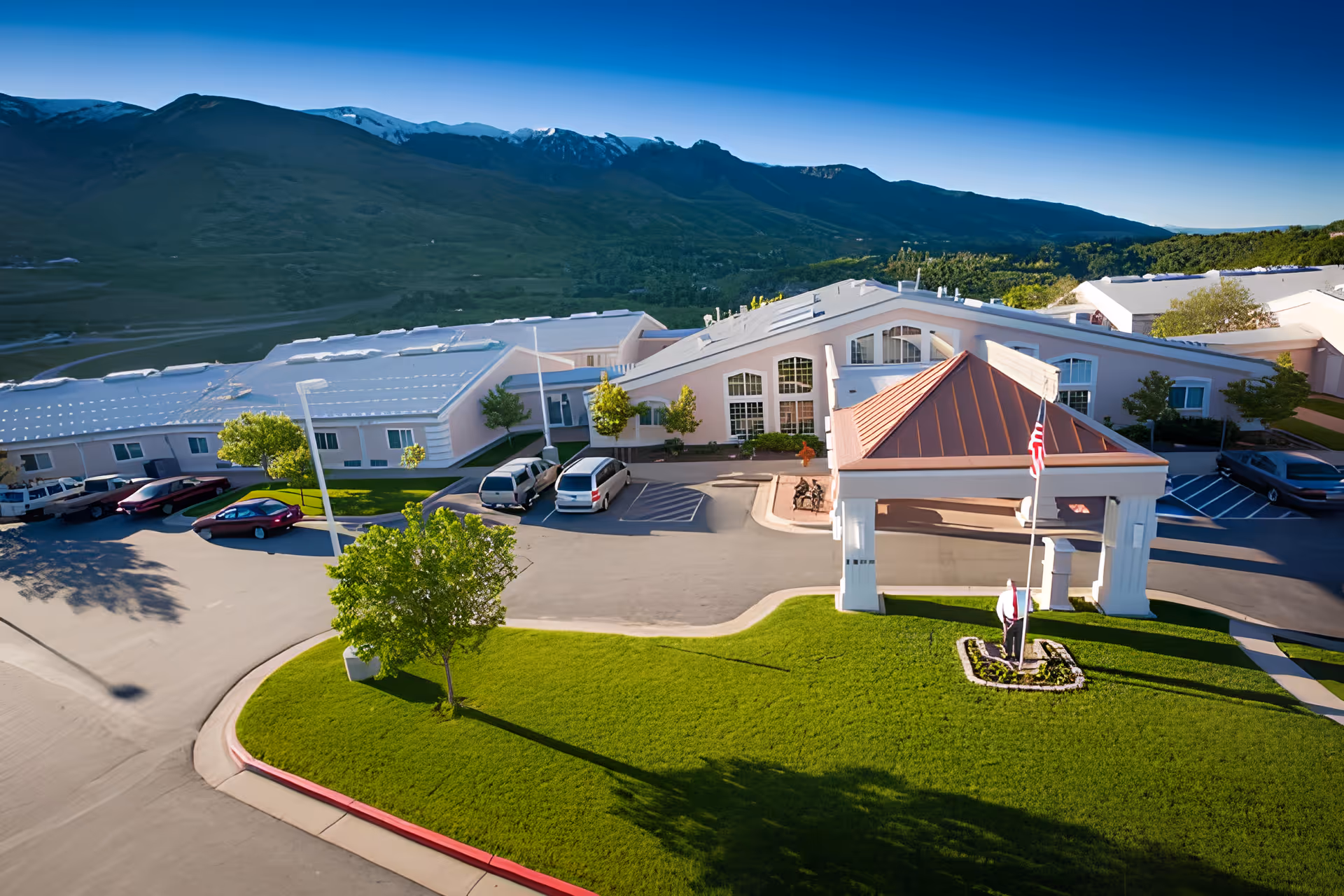 Aerial view of the Apple Inn At Apple Village facility showing a large building with a covered entrance and an American flag in front. Several cars are parked in the parking lot surrounding the building. The facility is set against a backdrop of green hills and mountains under a clear blue sky.