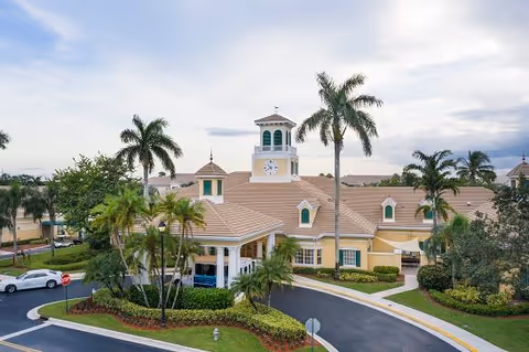 Front entrance of a Mediterranean-style senior living building with a clock tower, palm trees, and a circular driveway.