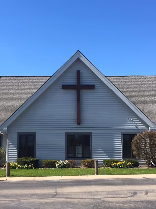 Front facade of a light-gray building with a large wooden cross mounted on the gable above three windows and landscaping.