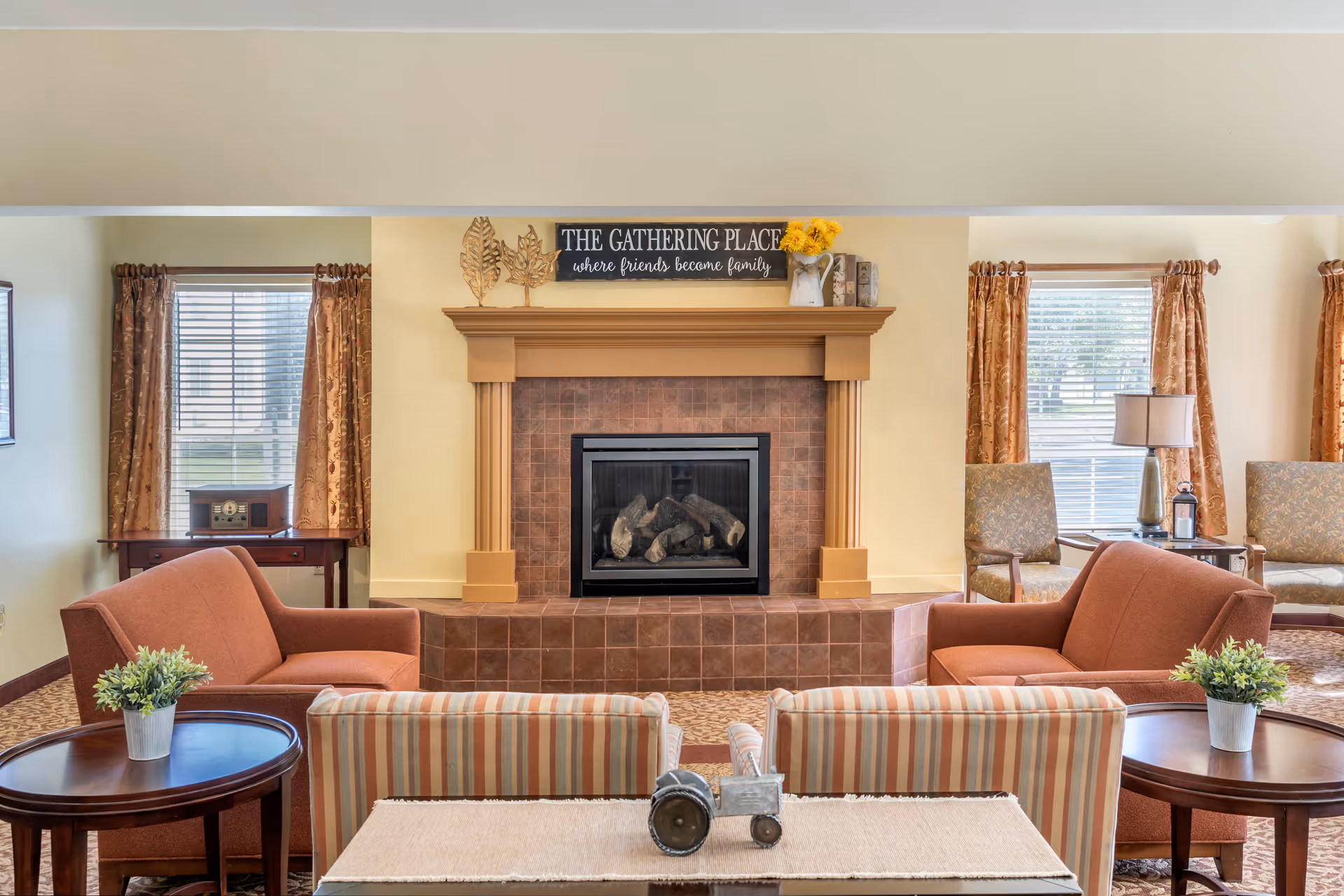 A cozy living room area with a central fireplace surrounded by brown tiles and a wooden mantle. Above the fireplace is a sign that reads 'THE GATHERING PLACE where friends become family.' The room features two orange armchairs facing each other, two striped armchairs facing the fireplace, and round wooden side tables with small potted plants. There are windows with patterned curtains on either side of the fireplace, and additional chairs and a lamp are visible in the background.