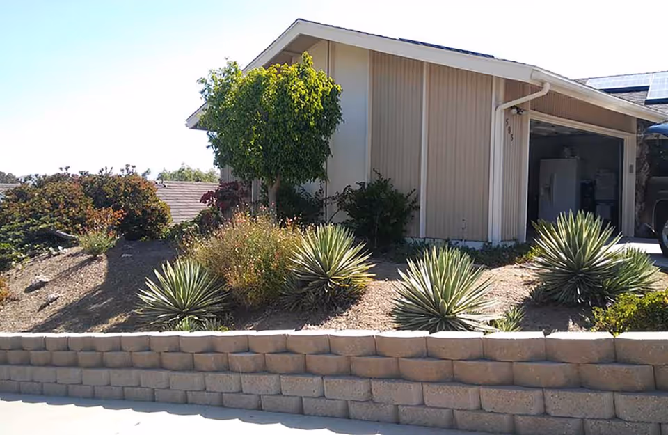Exterior view of a single-story building with beige siding and a sloped roof. The foreground features a landscaped area with various desert plants and shrubs bordered by a low retaining wall made of concrete blocks. The garage door is open, revealing a refrigerator and some stored items inside.