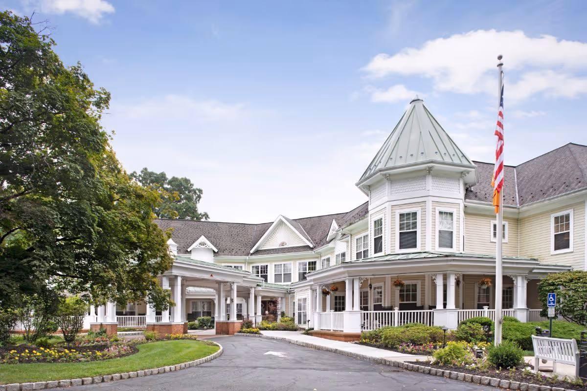 Front exterior of a large Victorian-style senior living facility with a wraparound porch, turret, American flag, driveway, and landscaped grounds.