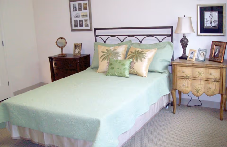 A neatly made bedroom with a green bedspread, palm-pattern pillows, and wooden nightstands.