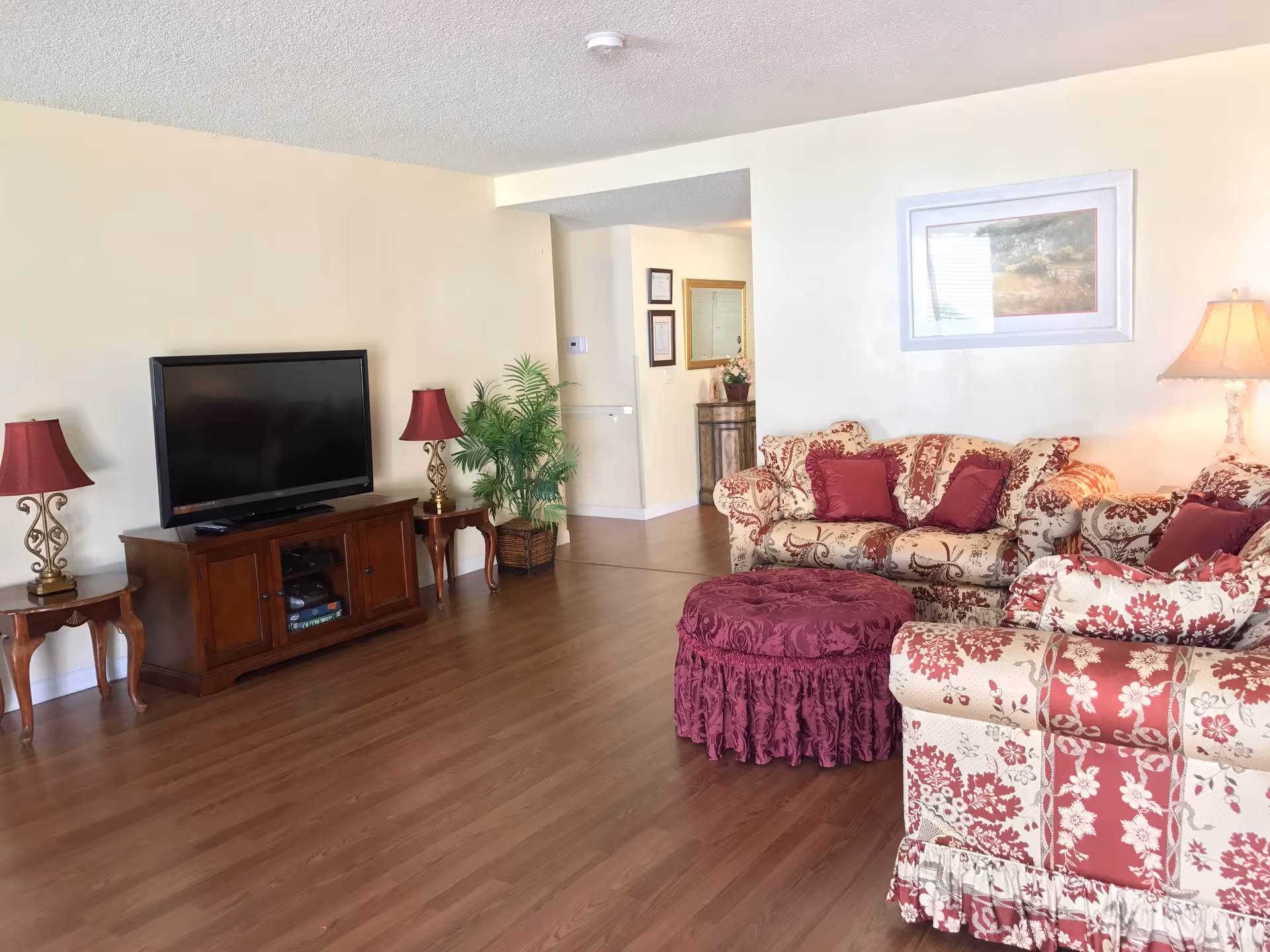 Spacious living room with floral sofa, ottoman, TV, and lamps in a care home.