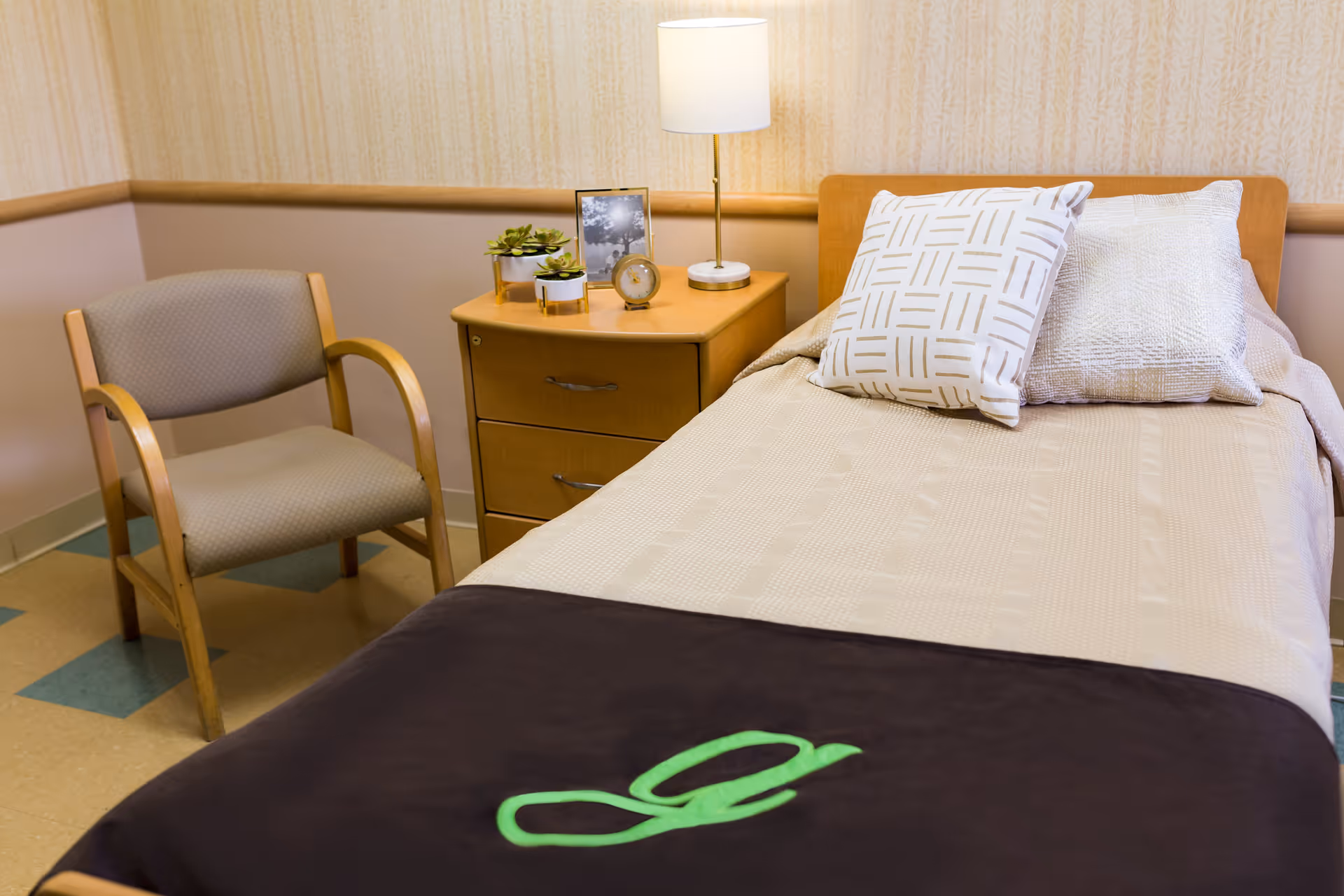 A neatly made single bed with two decorative pillows and a dark blanket featuring a green letter 'g' at the foot. Next to the bed is a wooden nightstand with a lamp, a small clock, two small potted plants, and a framed black and white photo. A wooden chair with cushioned seat and backrest is positioned beside the nightstand. The room has beige patterned wallpaper and a tiled floor with green and beige squares.