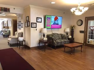 A cozy senior living facility lounge with couch, chairs, coffee table, lamps, and a wall TV.