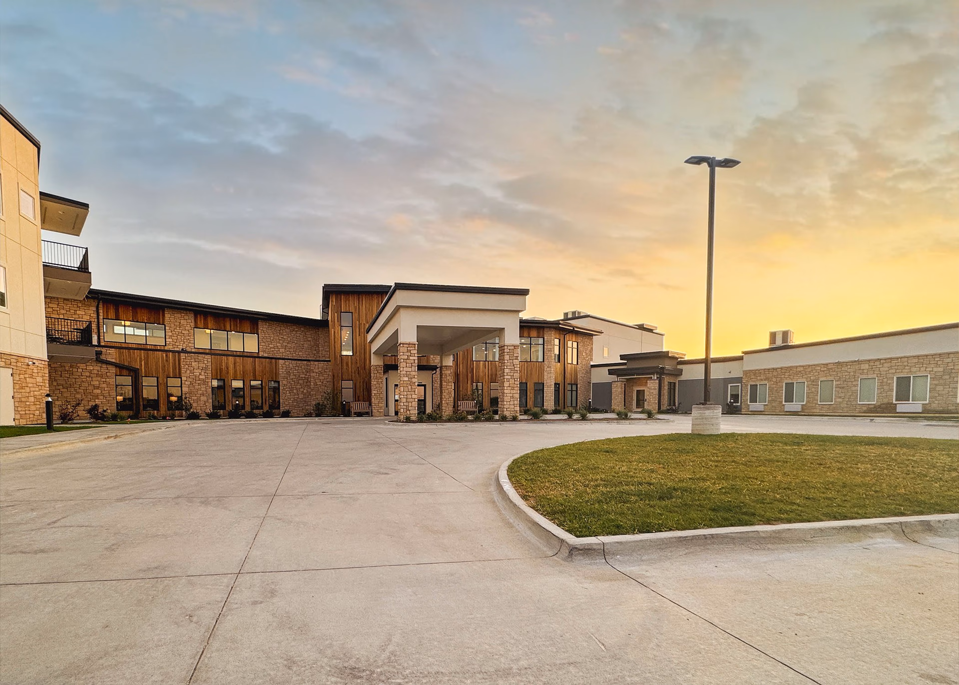 Front exterior of a senior living facility with a covered entrance, circular driveway, lawn, and a tall lamp post at sunset.