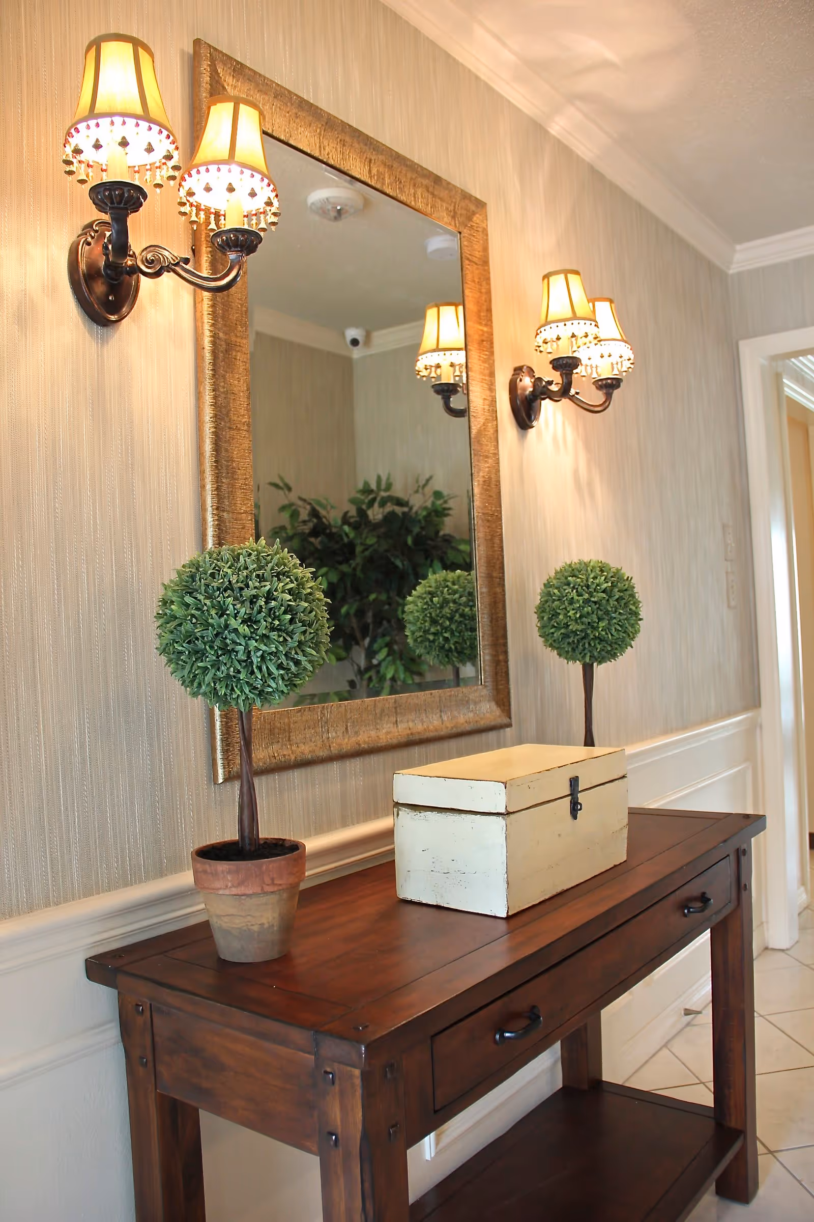 A wooden console table with two small potted topiary plants and a white decorative box on top. Above the table is a large framed mirror reflecting the opposite wall and a plant. Two wall-mounted lamps with beige lampshades flank the mirror, casting warm light on the textured wallpapered wall.