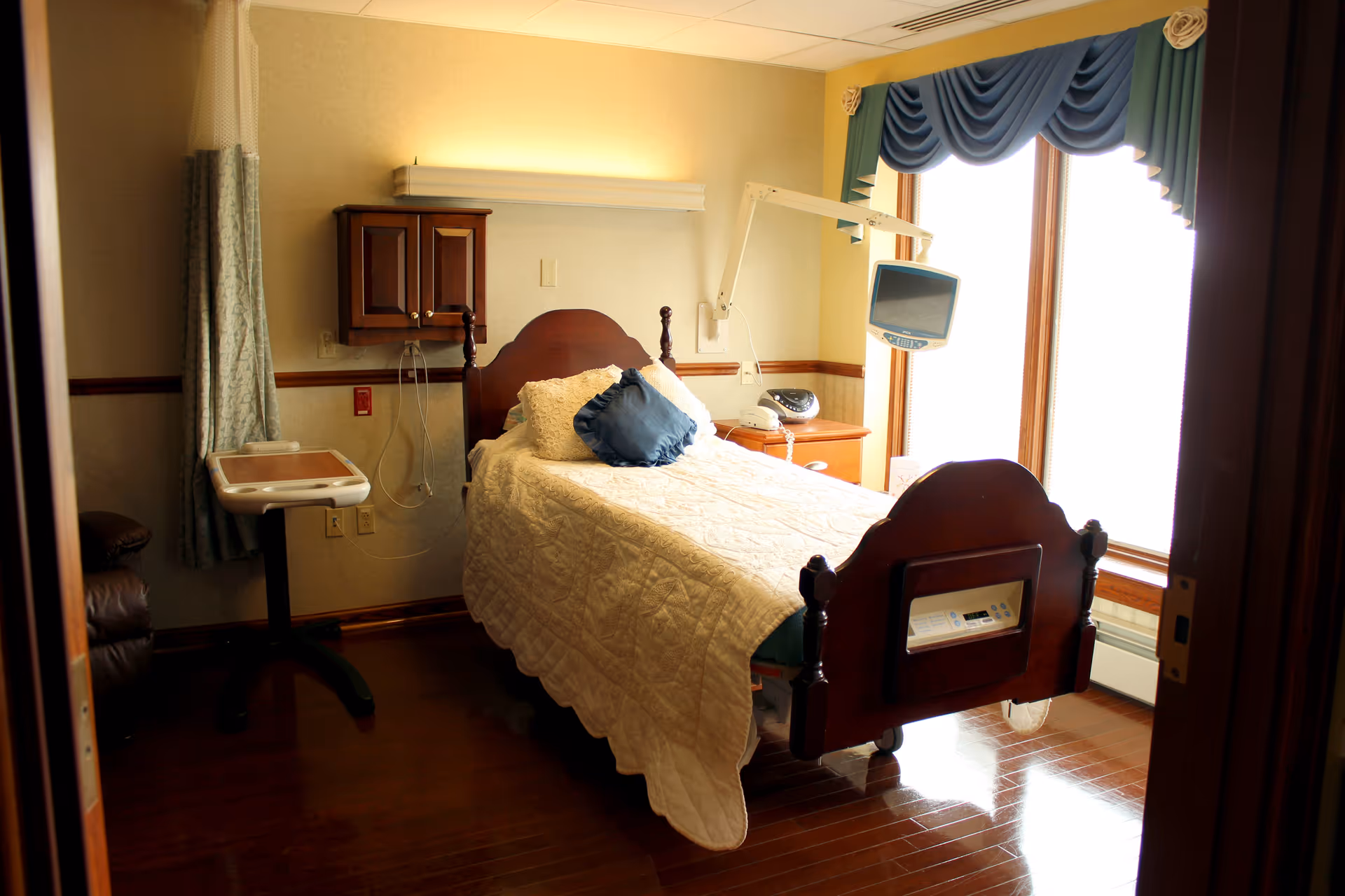 A hospice bedroom with a wooden hospital bed, monitor, and large window with curtains.