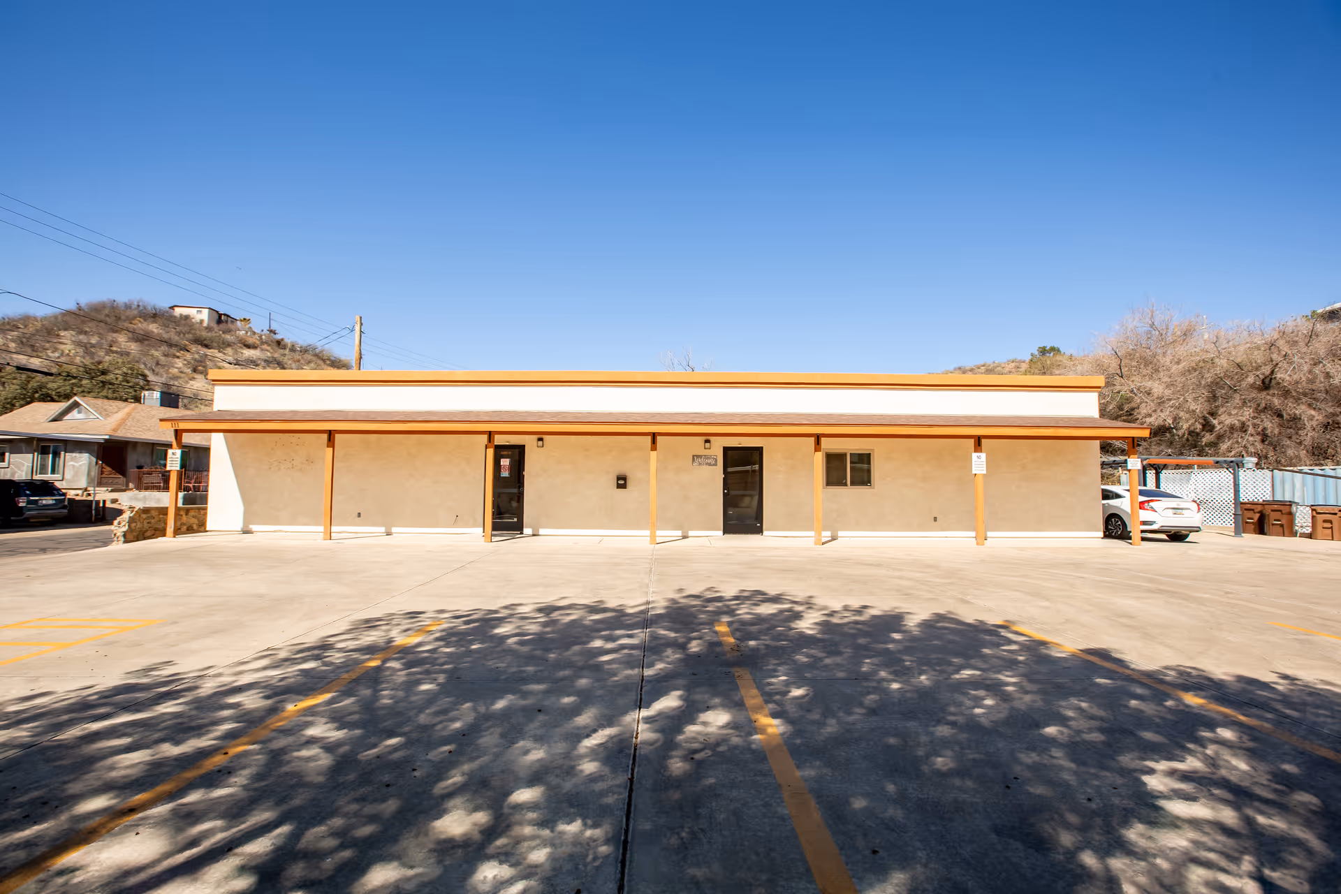 Single-story beige building with a wooden awning and empty parking lot in front.