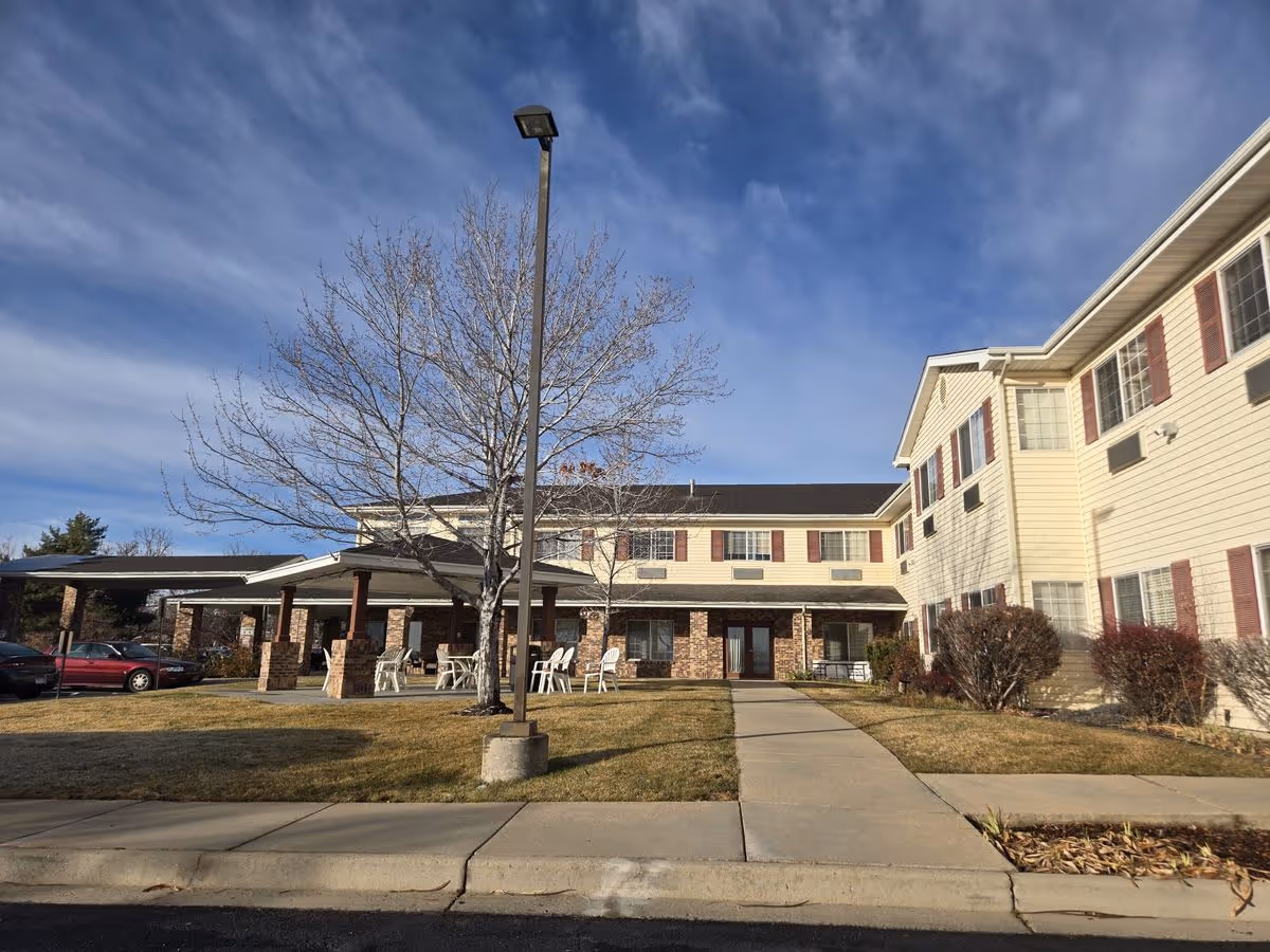Front exterior of a two-story senior living building with a covered entrance, lawn, and a leafless tree under a blue sky.