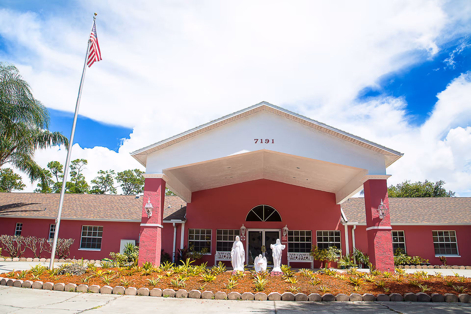Front entrance of a pink assisted living building with a covered portico displaying the number 7191, an American flag, statues, and landscaped flower beds.