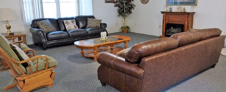 A cozy living room with two leather sofas, a wooden rocking chair with cushions, a wooden coffee table with a glass top, a floor lamp, a potted plant in the corner, and a wooden fireplace with decorative items on the mantel.