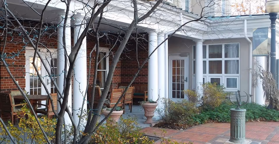 Covered brick porch with white columns and outdoor seating at a senior living entrance.