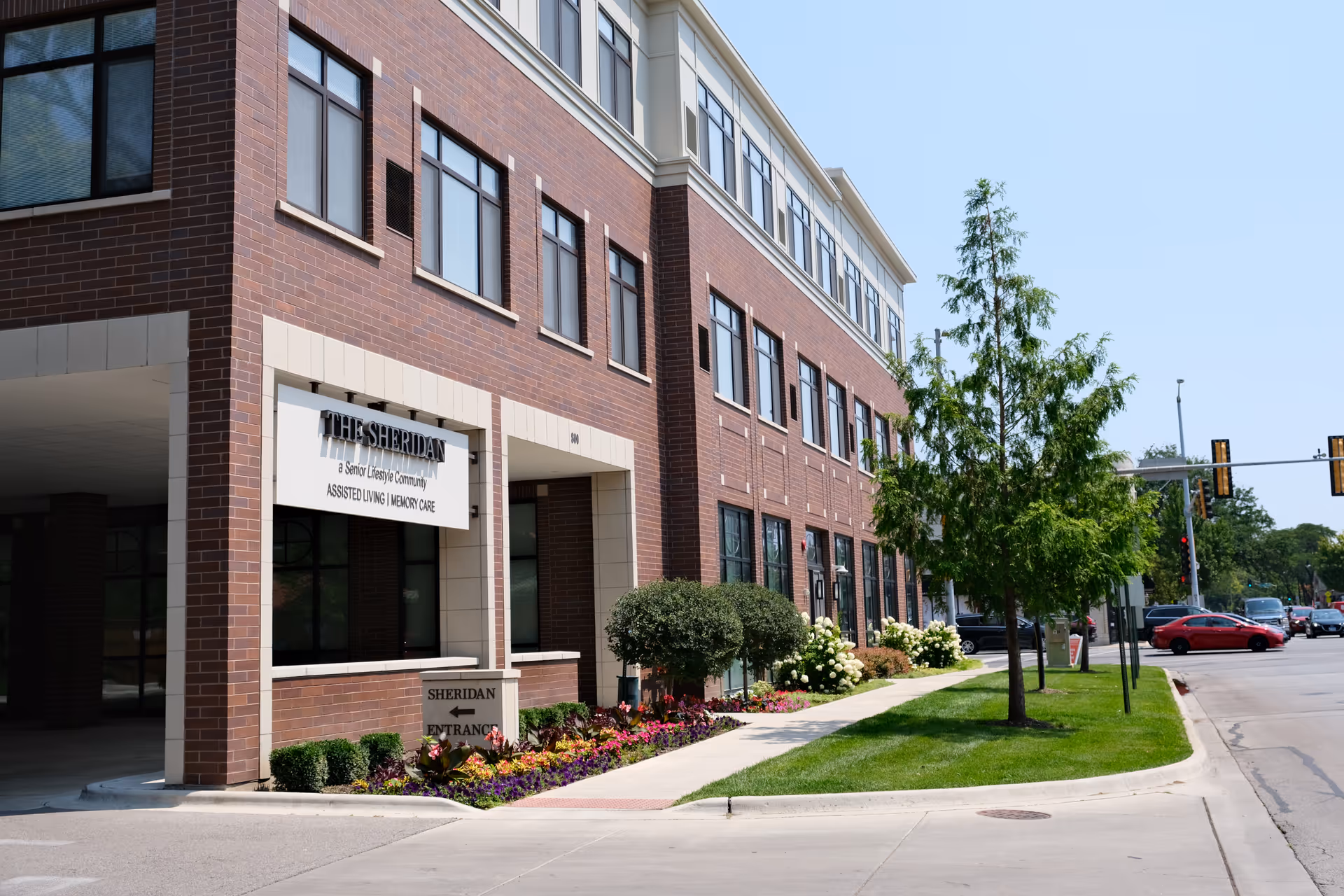 Exterior view of The Sheridan at River Forest building, a senior lifestyle community offering assisted living and memory care. The building is made of red brick with multiple windows, a landscaped sidewalk with flowers and bushes, and a tree near the street corner.