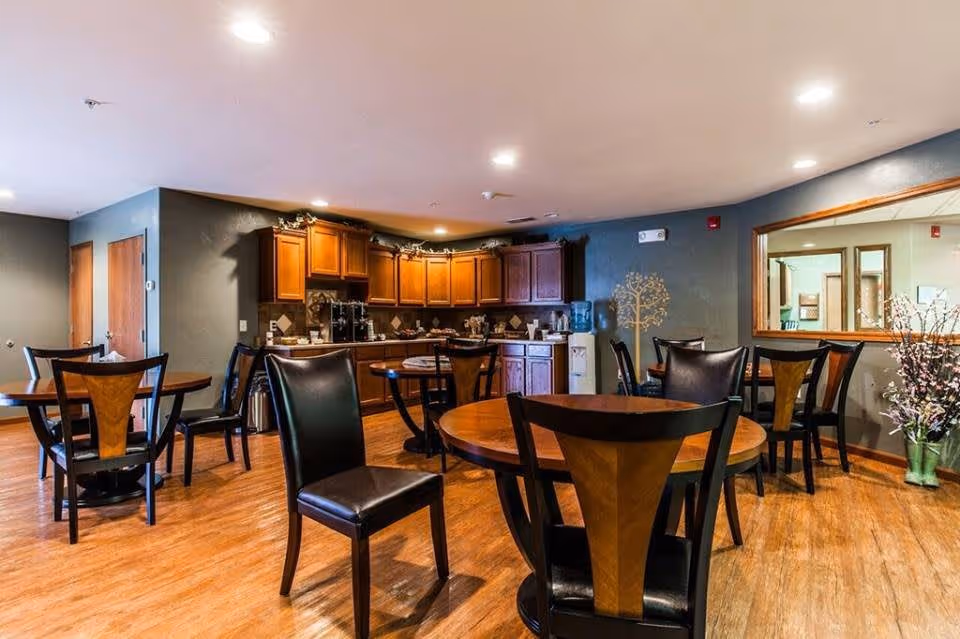 A dining area with several round wooden tables and black chairs with wooden backs. The room has wooden flooring and a kitchen area with wooden cabinets and a countertop in the background. There is a water dispenser and decorative plants near a large window on the right wall.
