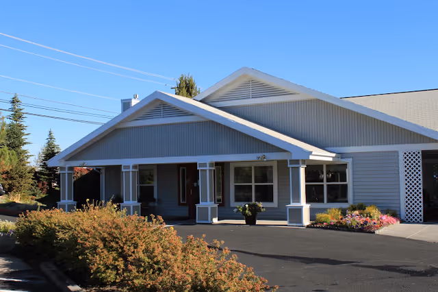 Exterior view of a single-story building with a covered entrance supported by white columns. The building is light gray with white trim and surrounded by bushes and flowers under a clear blue sky.