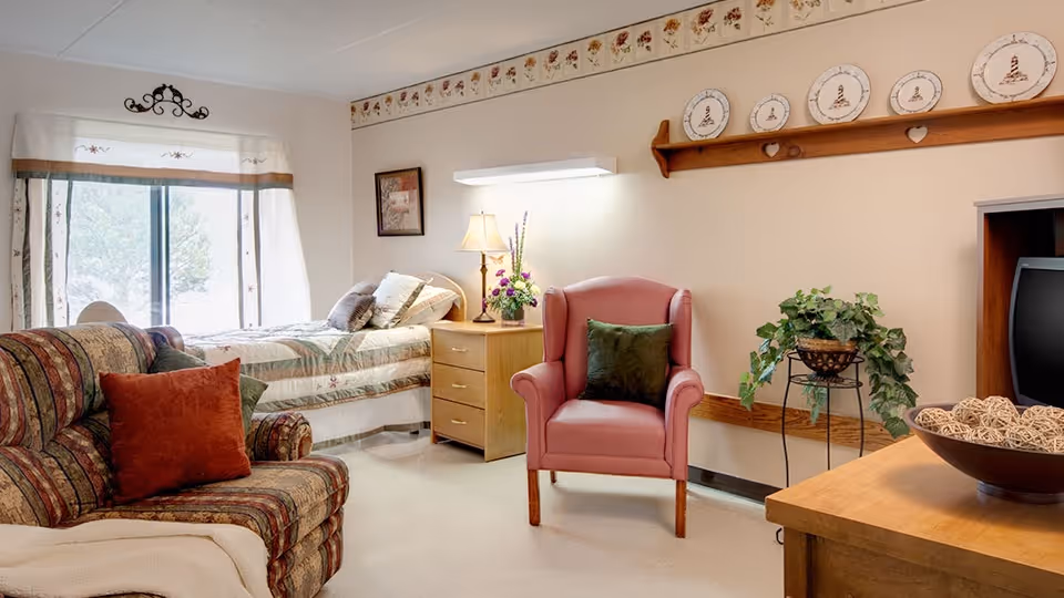 Sunlit senior living bedroom with a bed, armchair, sofa, side table with a lamp, and decorative plates on a wall shelf.