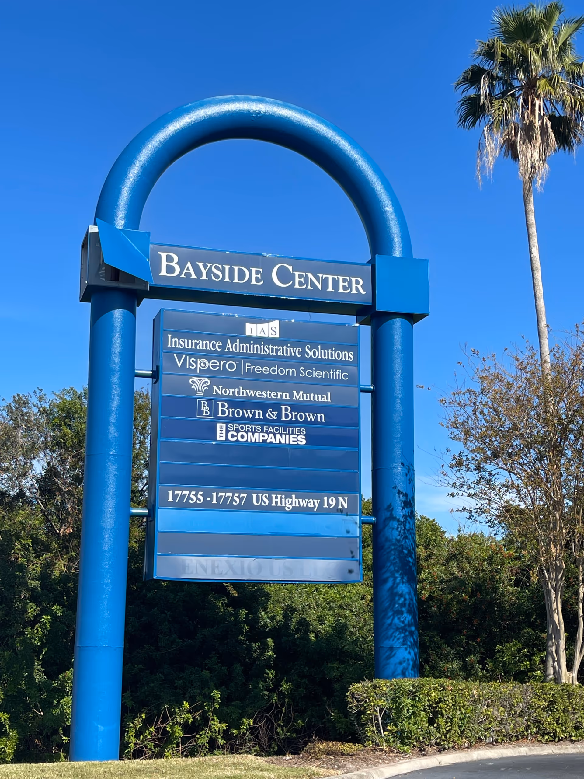 Blue arch sign reading Bayside Center with listings, outdoors beside greenery.
