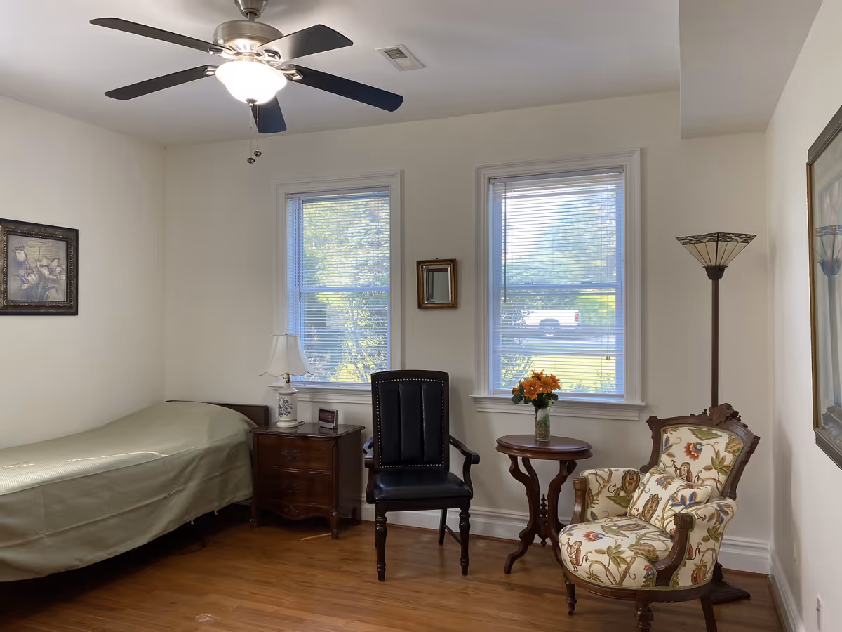 A small bedroom with a single bed covered in a green bedspread, a wooden nightstand with a lamp and clock, two windows with blinds, a black leather chair, a round wooden side table with a vase of orange flowers, a floral upholstered armchair, a floor lamp, and framed artwork on the walls.