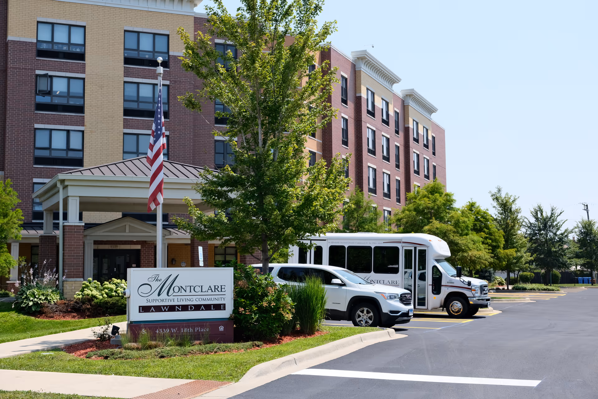 Exterior view of The Montclare Supportive Living Community building with its entrance sign, parked shuttle and car, flagpole, and landscaped grounds.