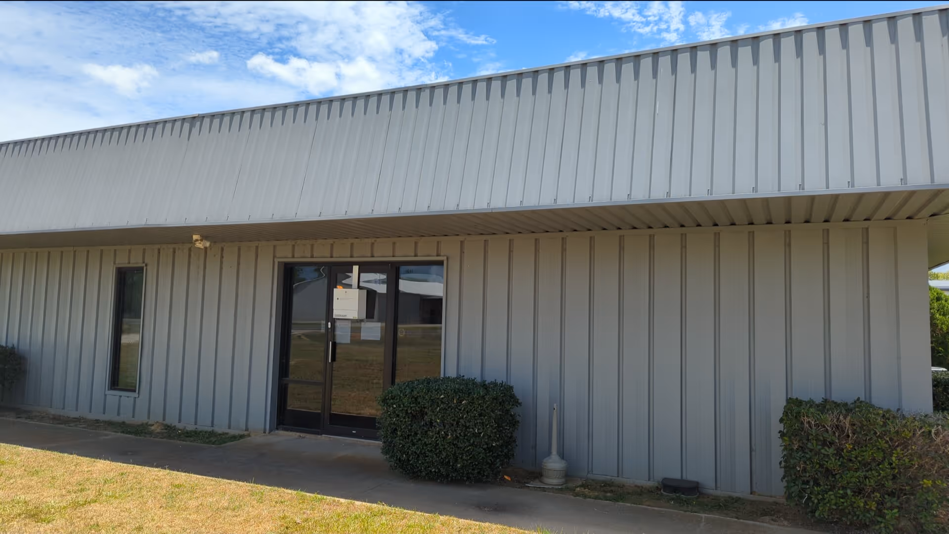 Gray metal building exterior with glass entrance doors and trimmed shrubs.