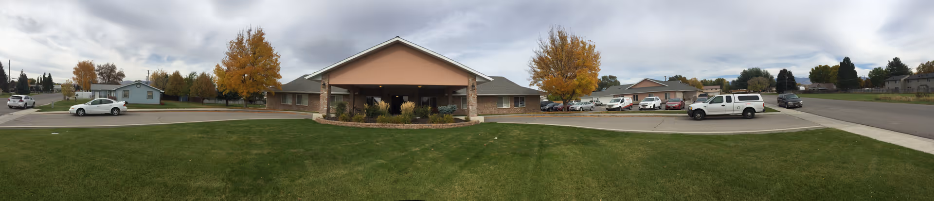 Wide panoramic view of the front exterior of Heritage Senior Living facility showing a single-story building with a covered entrance, surrounded by a well-maintained lawn and several parked cars. Trees with autumn foliage are visible around the building under a cloudy sky.