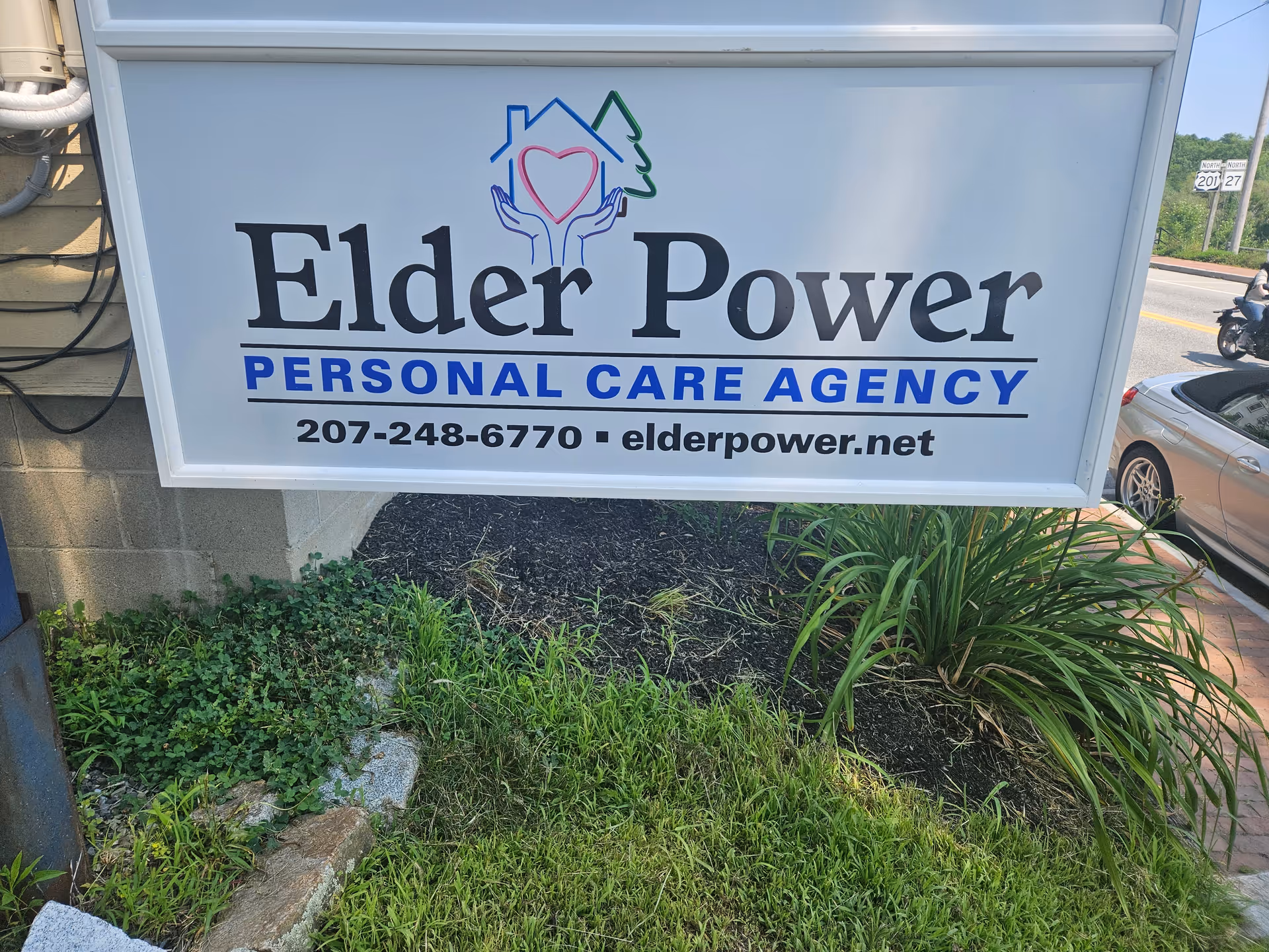 Exterior sign for Elder Power Personal Care Agency with phone number and website amid landscaping.