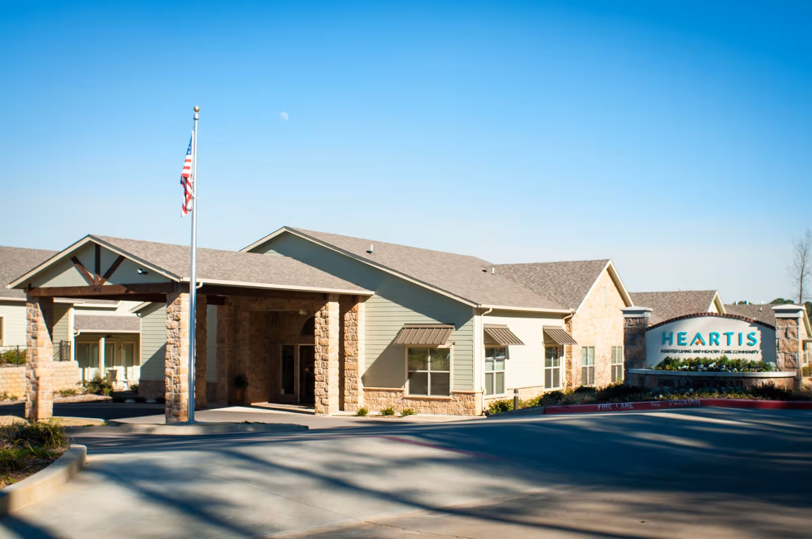 Exterior view of Heartis Longview assisted living and memory care community building with a covered entrance supported by stone pillars, an American flag on a flagpole, and a clear blue sky.
