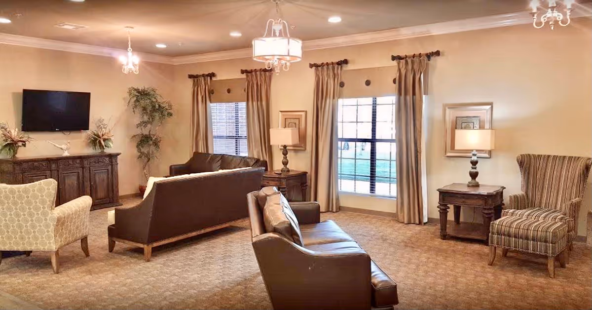 A spacious living room with beige walls and carpeted floor, featuring two brown leather sofas, a patterned armchair, a striped wingback chair, wooden side tables with lamps, a large window with beige curtains, a flat-screen TV mounted on the wall above a wooden cabinet, and decorative plants.