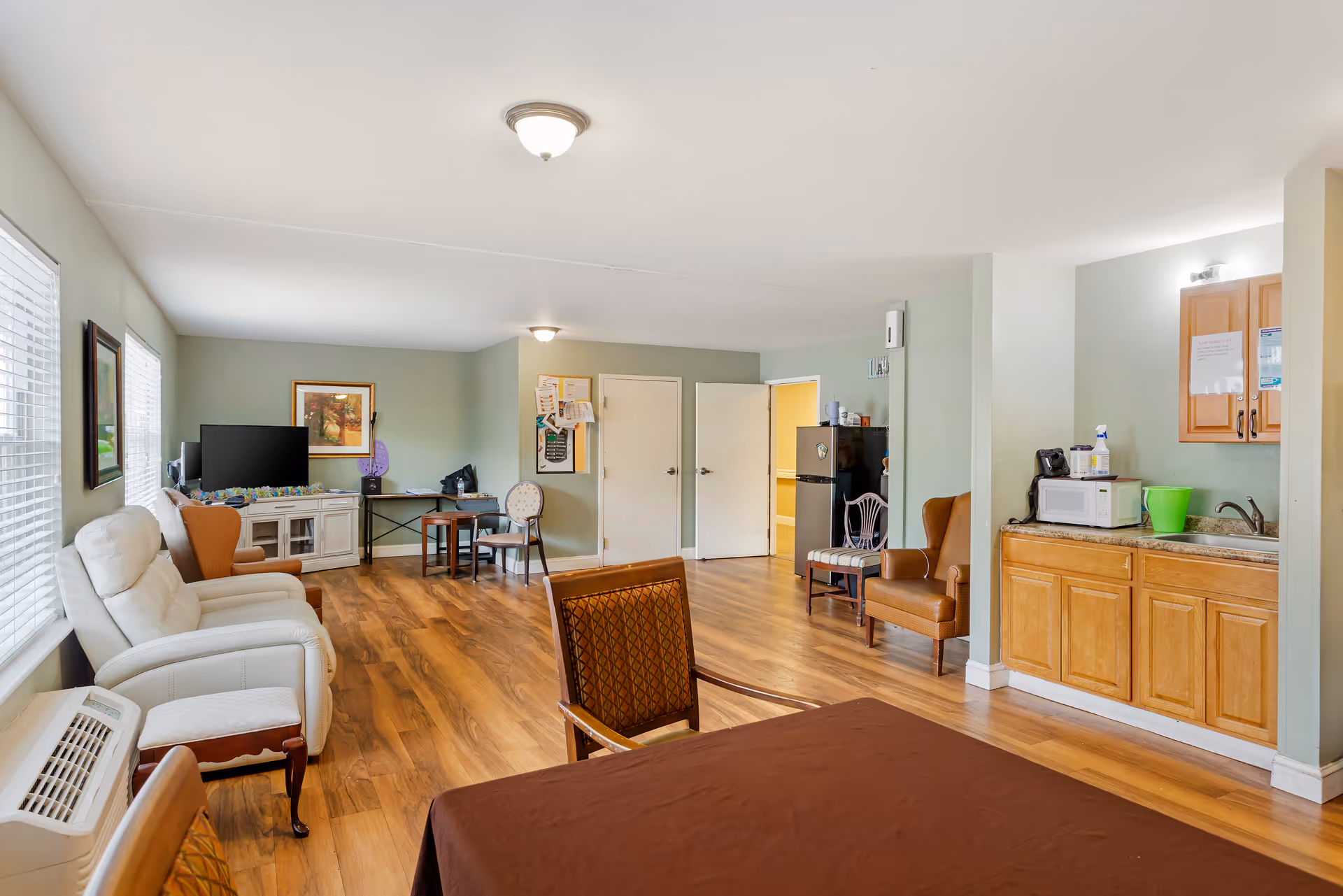 Spacious senior living common area with seating, tables, and a kitchenette along light green walls.