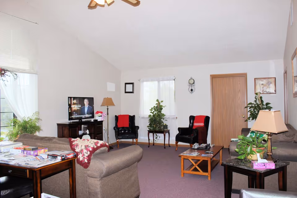 Cozy senior living lounge with sofas, chairs, plants, and a TV in a common area.