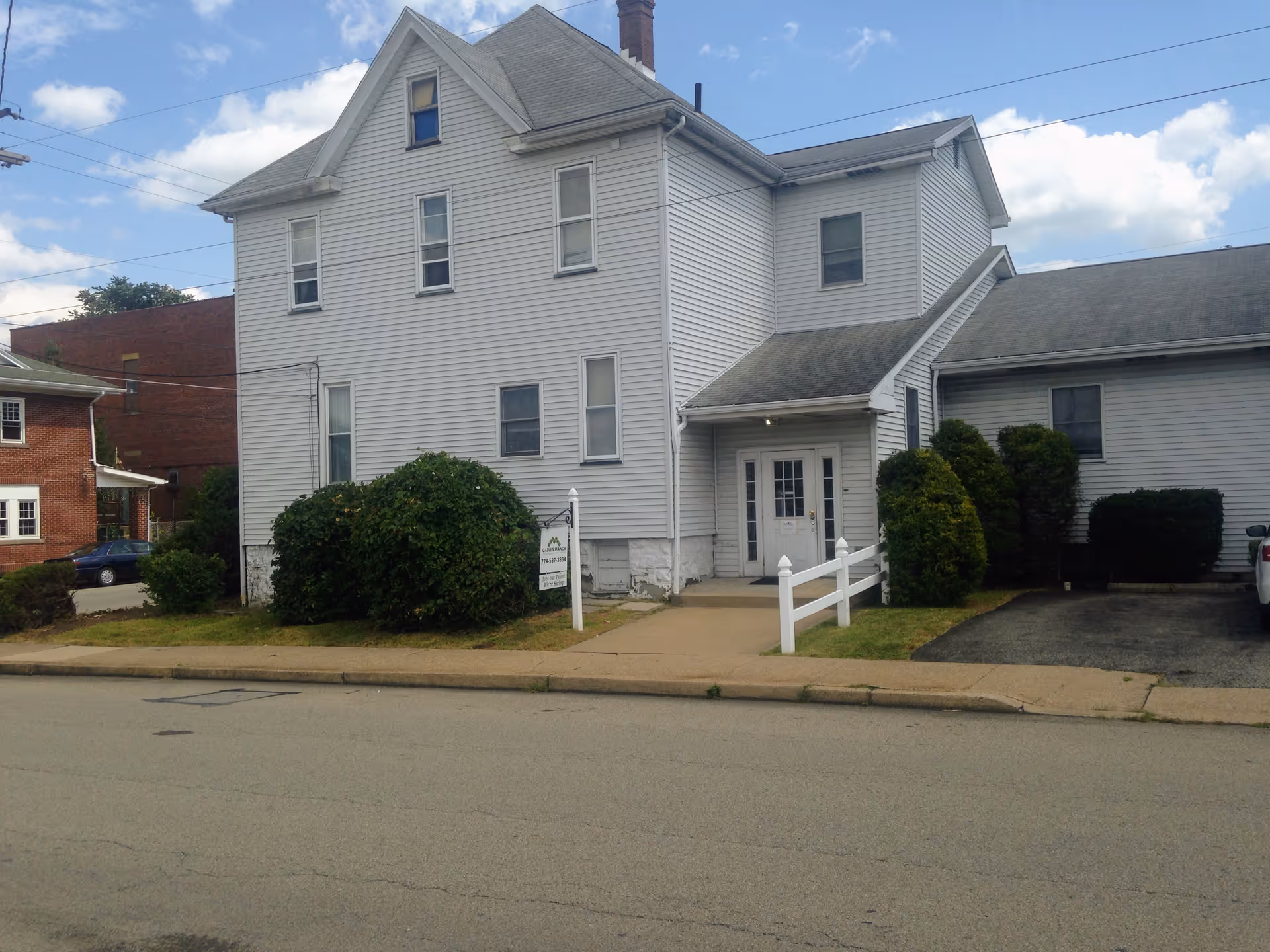 Exterior view of a two-story white building with multiple windows and a small entrance porch. There are bushes and a small lawn in front, along with a sidewalk and a driveway. The sky is partly cloudy.
