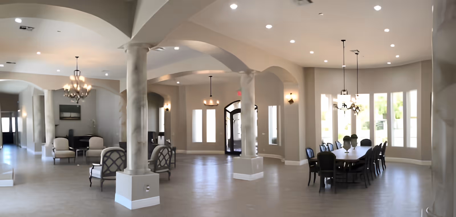 Spacious and well-lit interior of a senior living facility featuring a large dining table with chairs on the right side, several seating areas with armchairs on the left, decorative columns, arched doorways, and multiple ceiling lights and chandeliers.