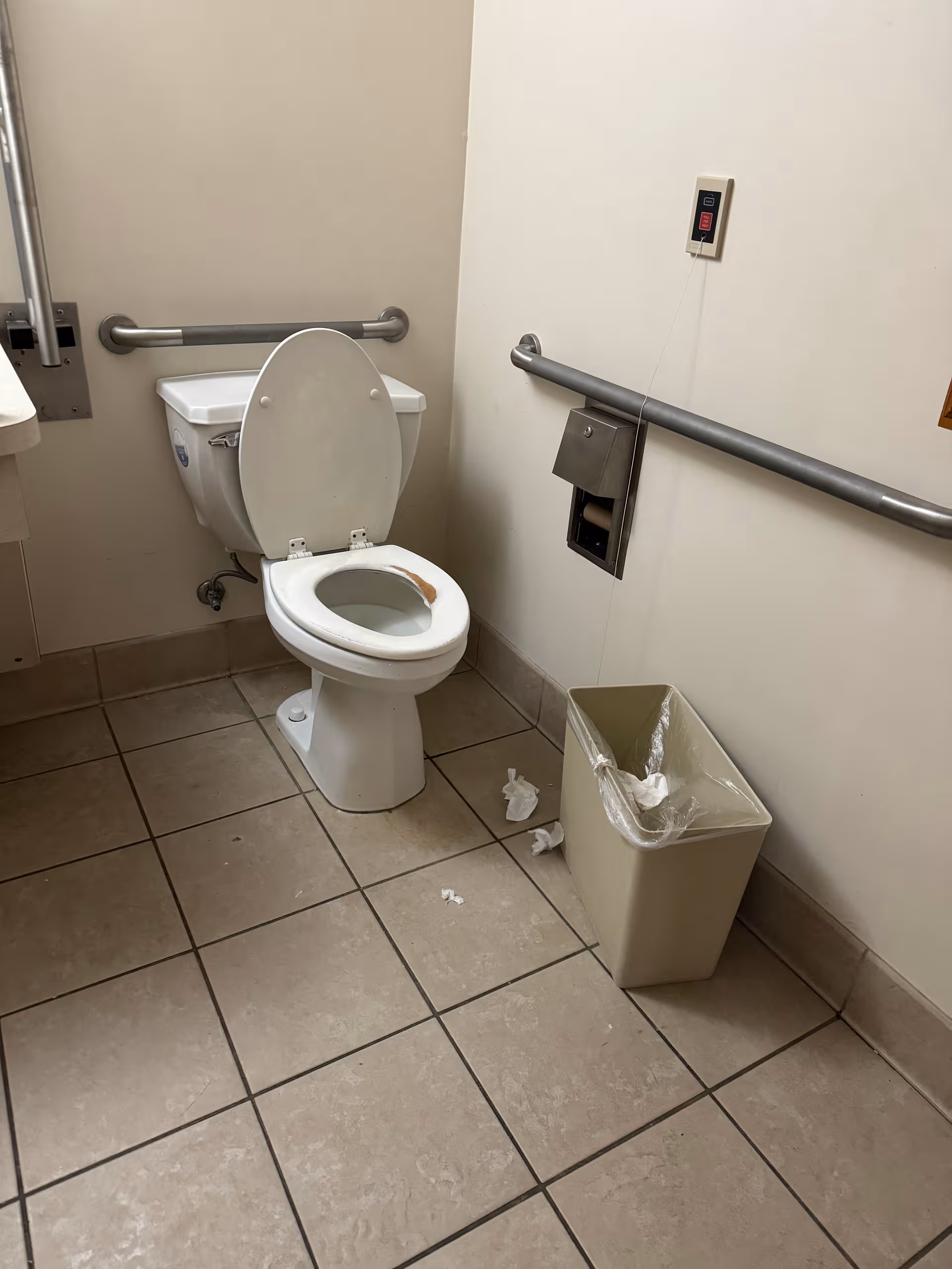 Accessible restroom with a toilet, grab bars, and a trash bin in a tiled room.