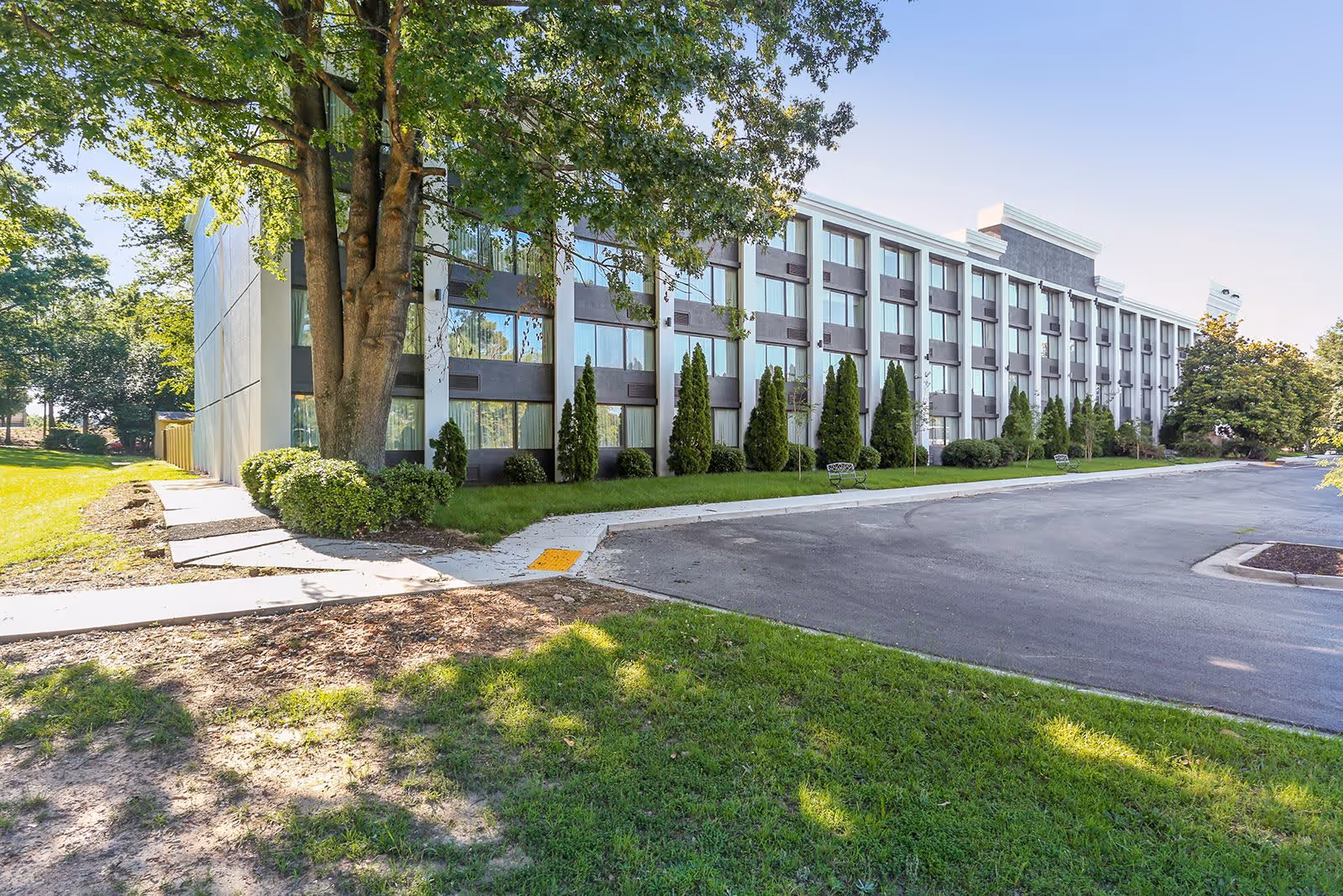 Exterior view of a multi-story senior living facility building with large windows, surrounded by green grass, trees, and shrubs under a clear blue sky.