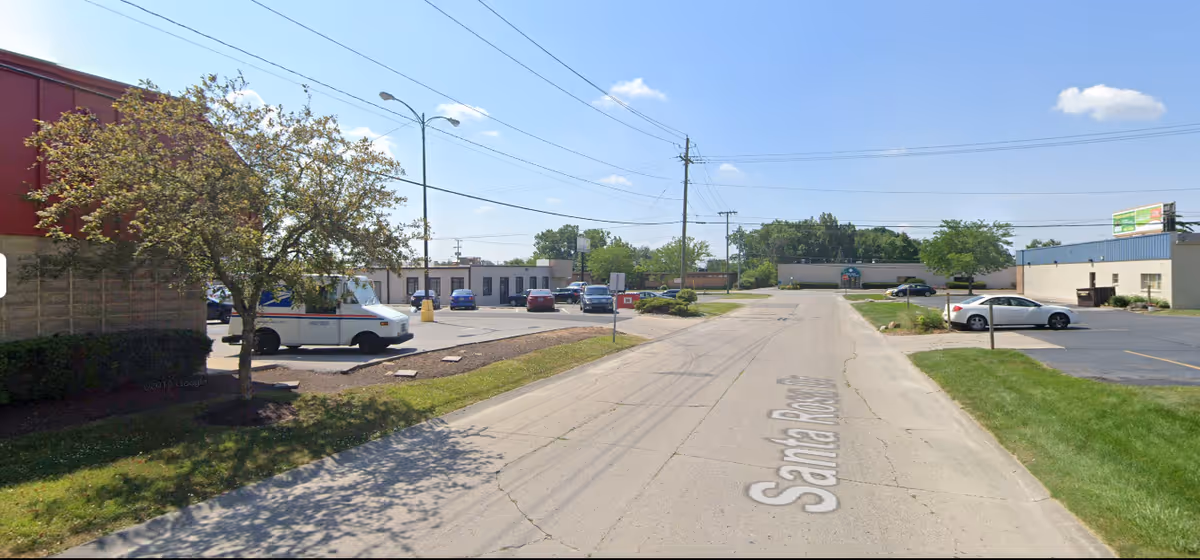 Outdoor street scene with parked cars, trees, and low-rise buildings along a road.