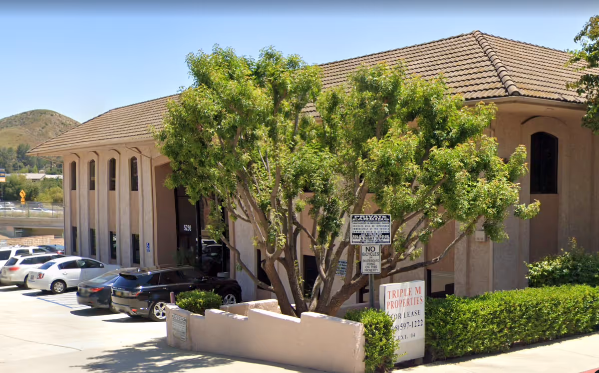 Exterior view of a senior living facility with a tan tiled roof, parked cars, and trees.