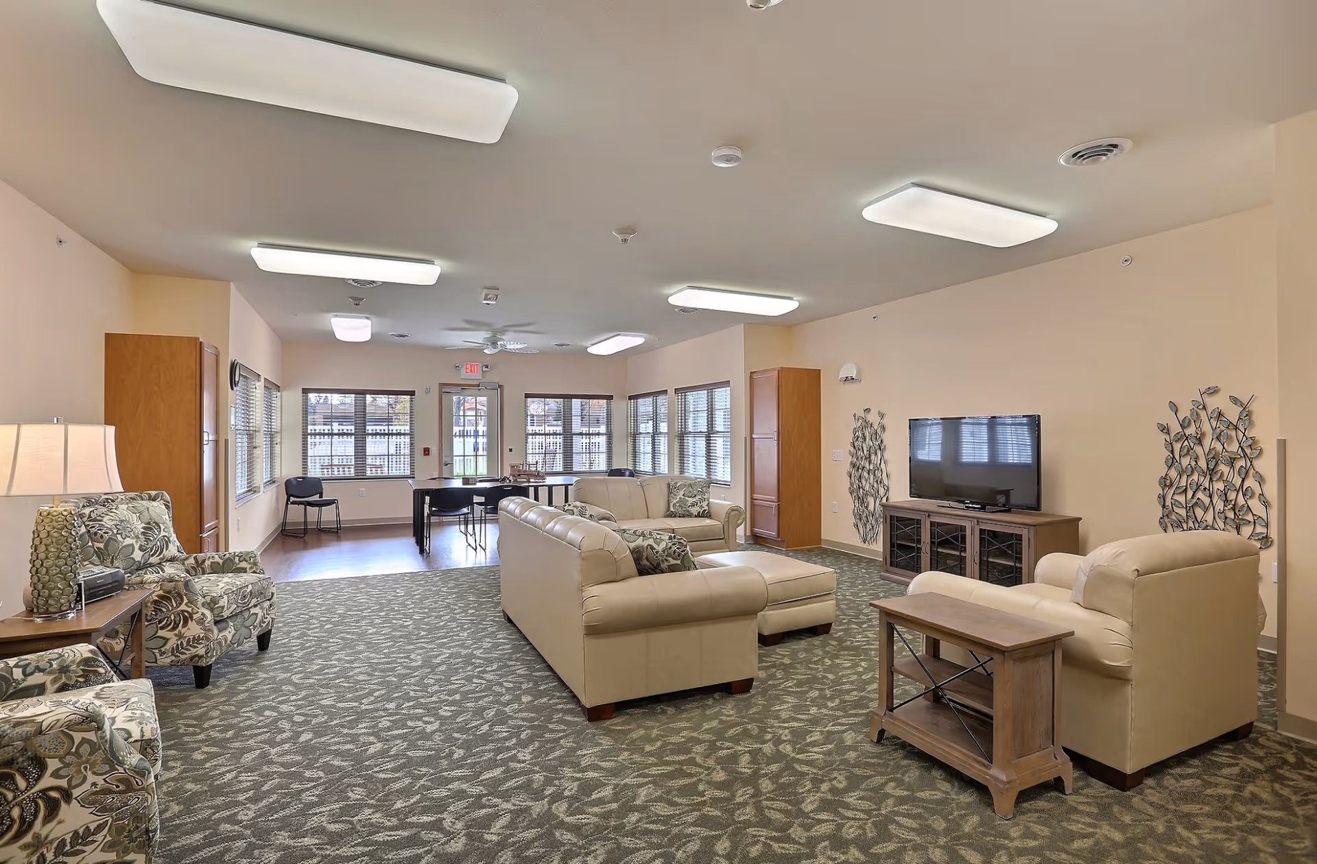 Spacious communal lounge with beige sofas, patterned armchairs, a TV console, and large windows.