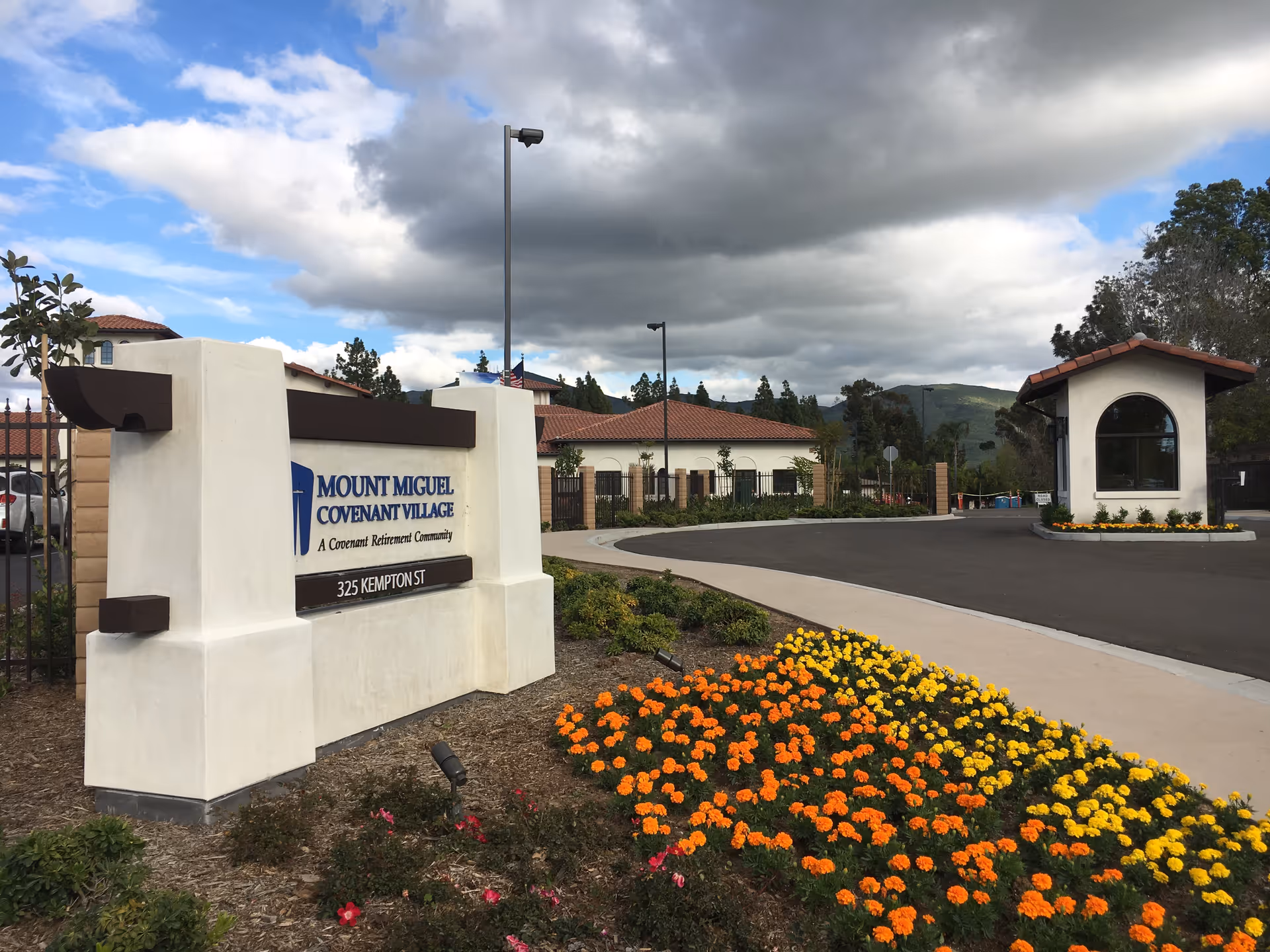 Entrance to Covenant Village Mount Miguel with a sign, gatehouse, and colorful flower beds.