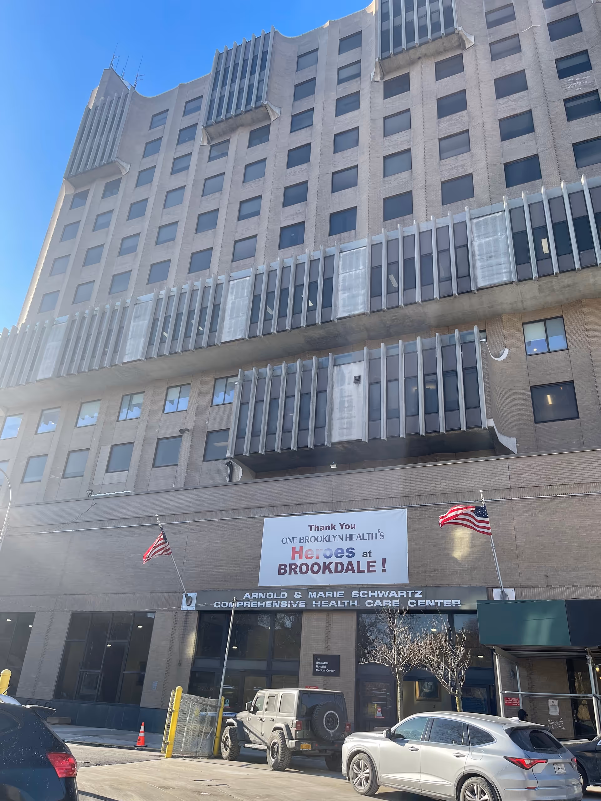 Exterior of One Brooklyn Health-Brookdale facility with banners, entrance, and parked cars.