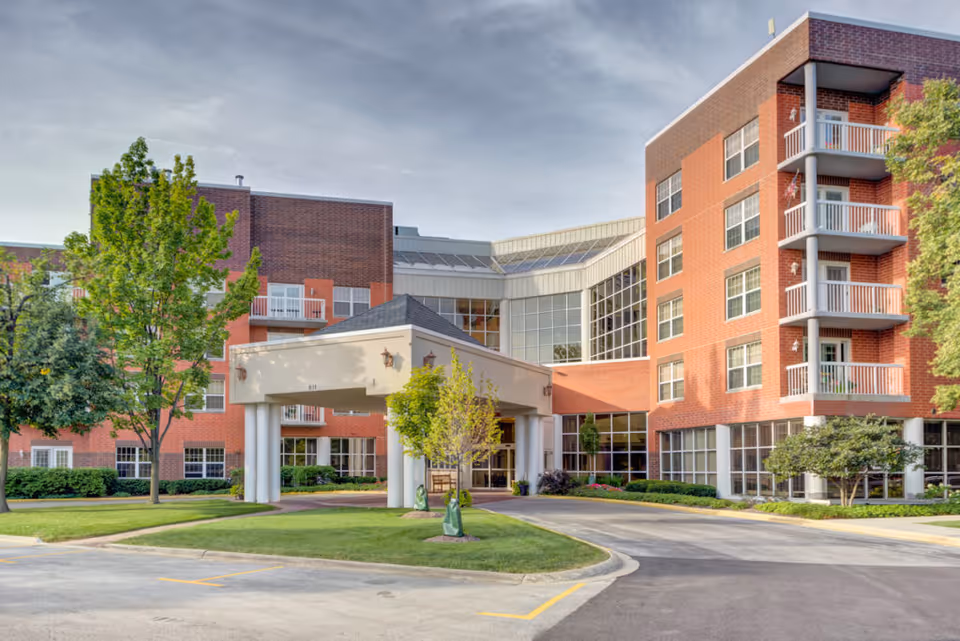 Exterior front entrance of a multi-story red-brick senior living building with a covered porte-cochère, balconies, and landscaped grounds.