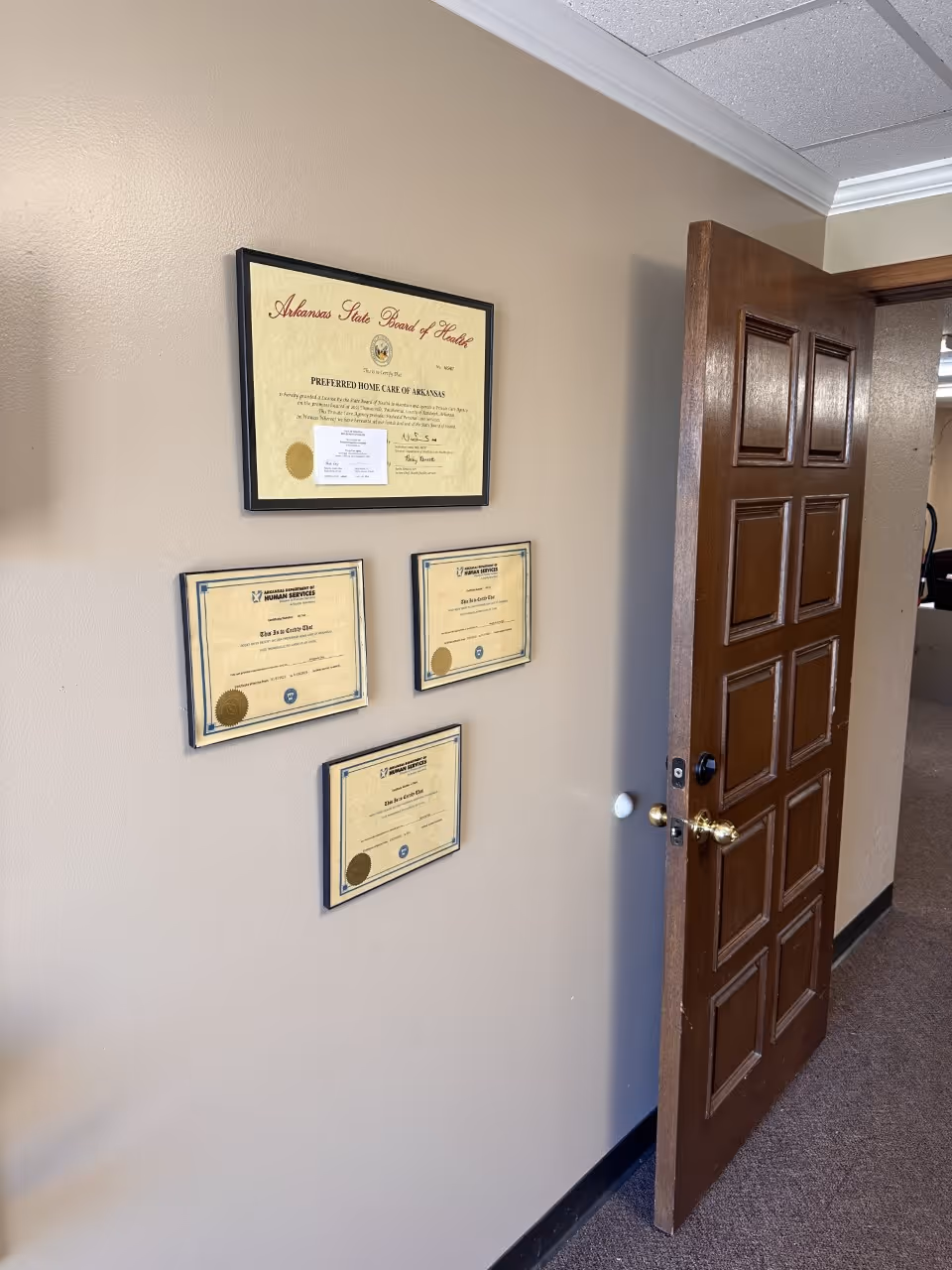 Brown door ajar in a beige hallway with framed certificates on the wall.