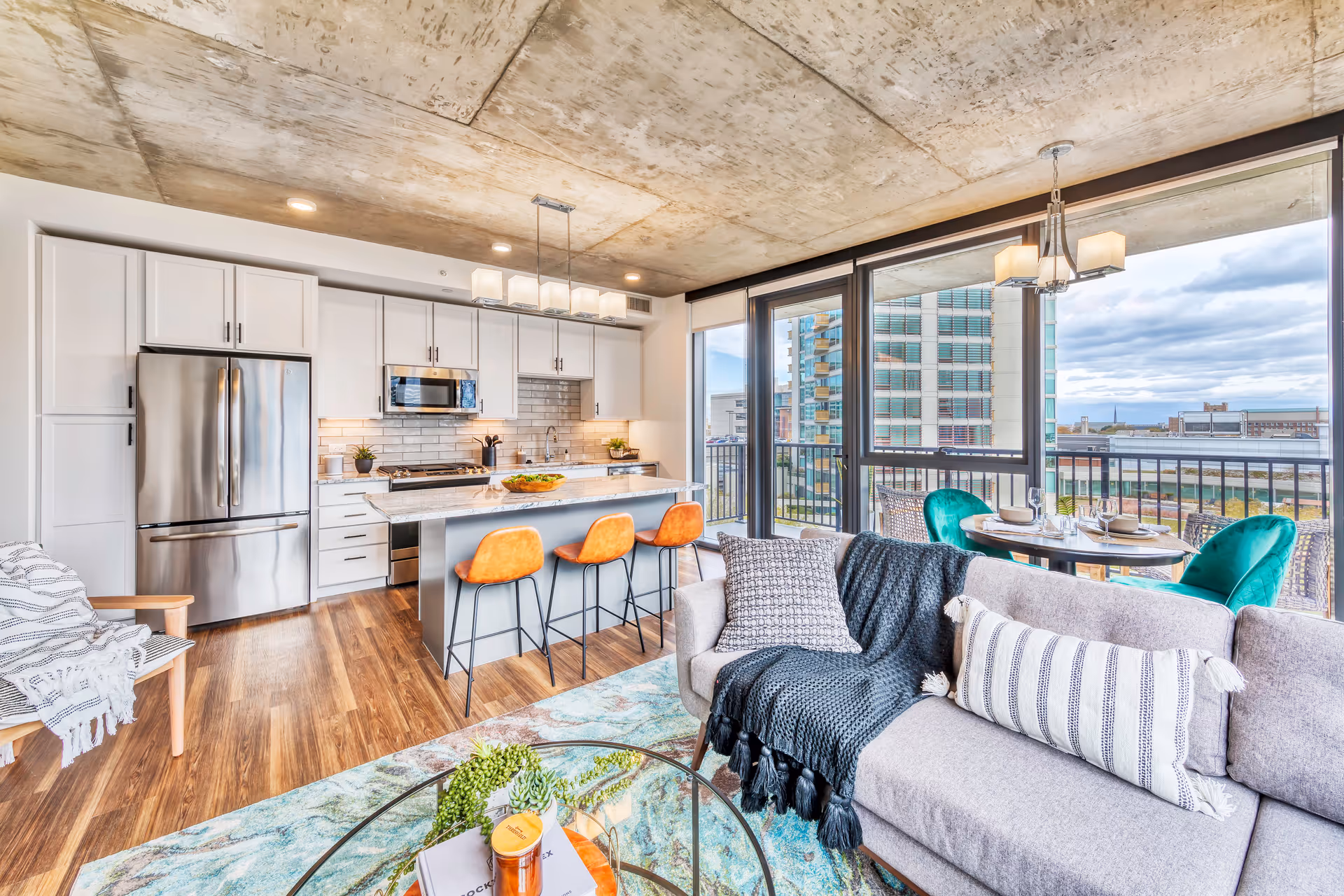 Bright open-concept living room and kitchen with an island and orange bar stools, large windows overlooking a balcony and city view.