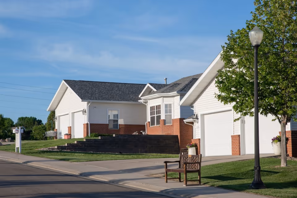 Single-story white and brick residential building with garages, a streetlamp, tree, and bench along a sidewalk under a blue sky.