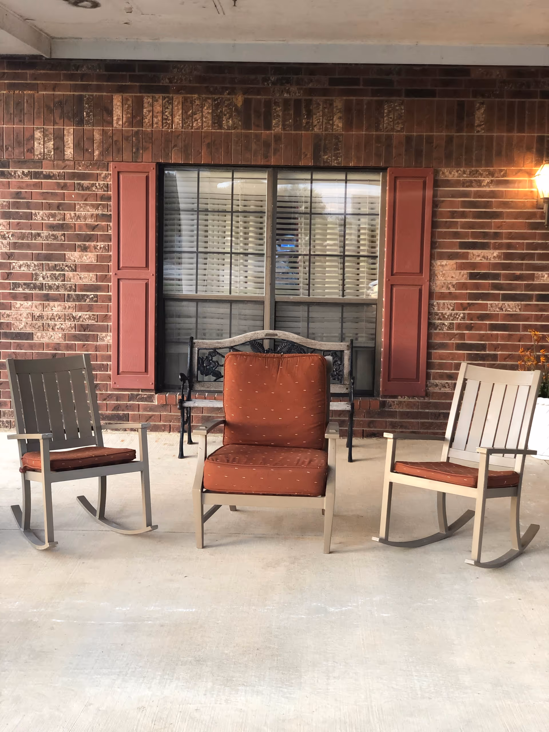 Outdoor covered porch with three cushioned rocking chairs and a brick wall behind.
