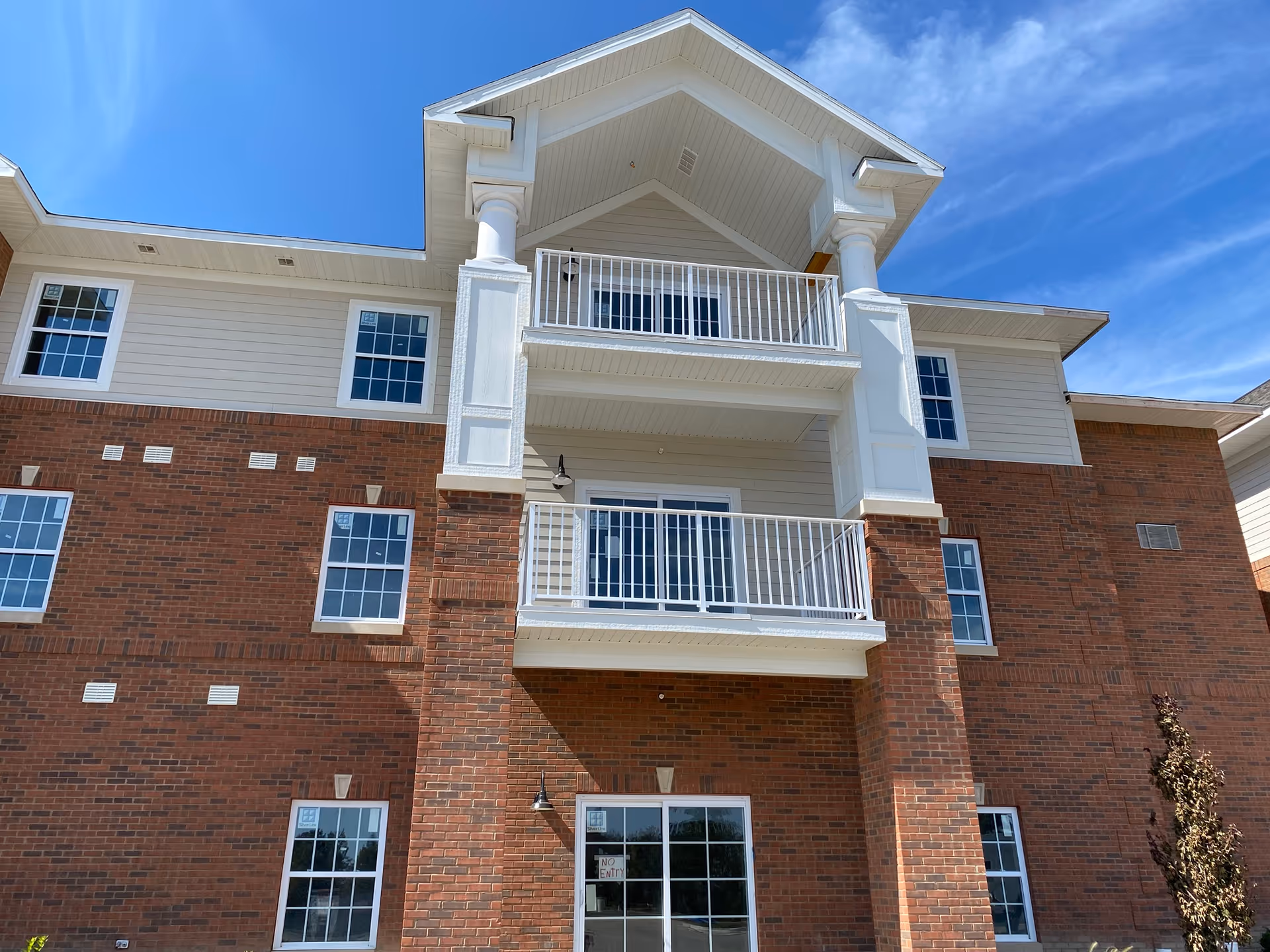 Front exterior of a three-story brick and siding building with white balconies and railings under a blue sky.