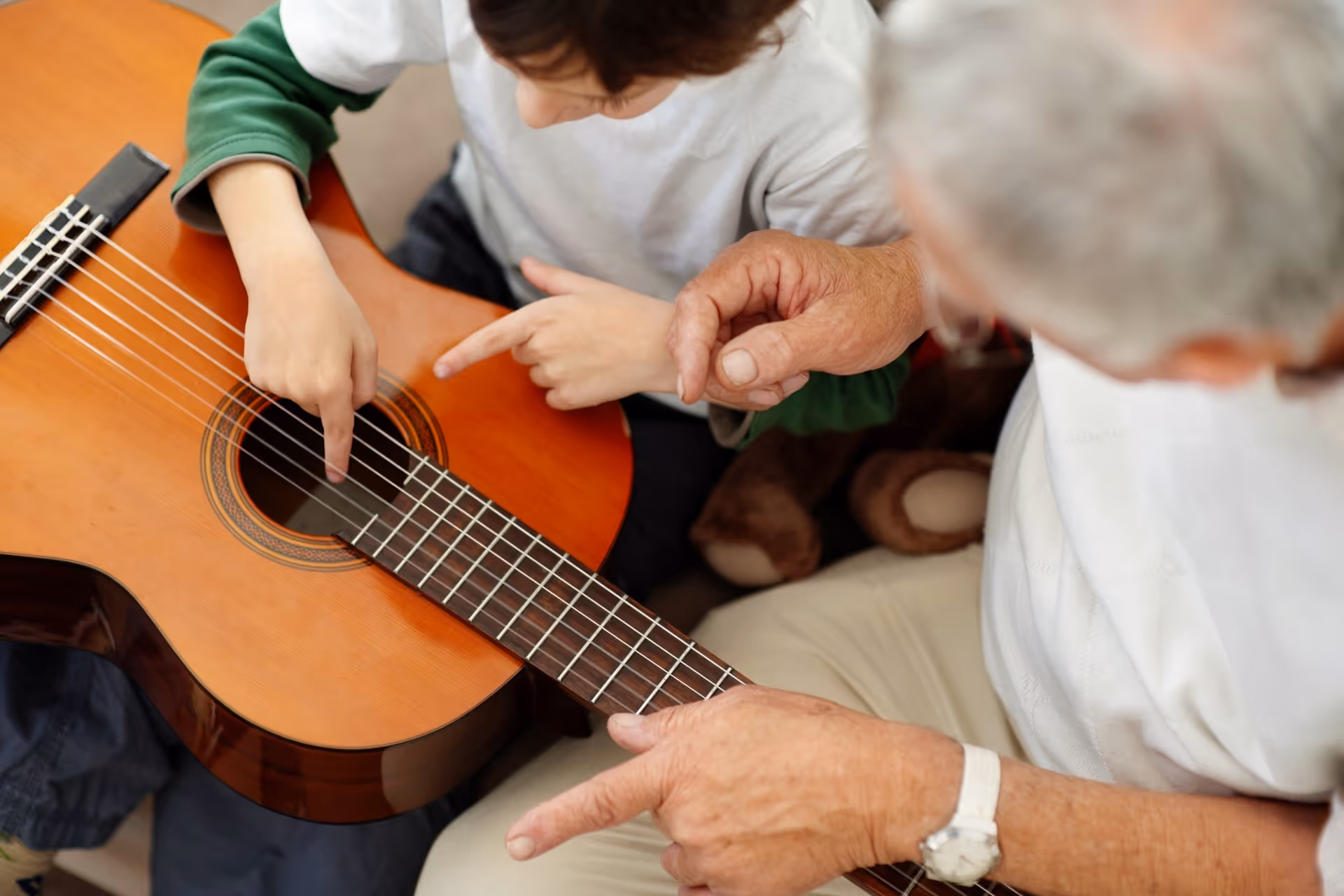 Two people interact indoors, playing a guitar with one pointing toward the strings.