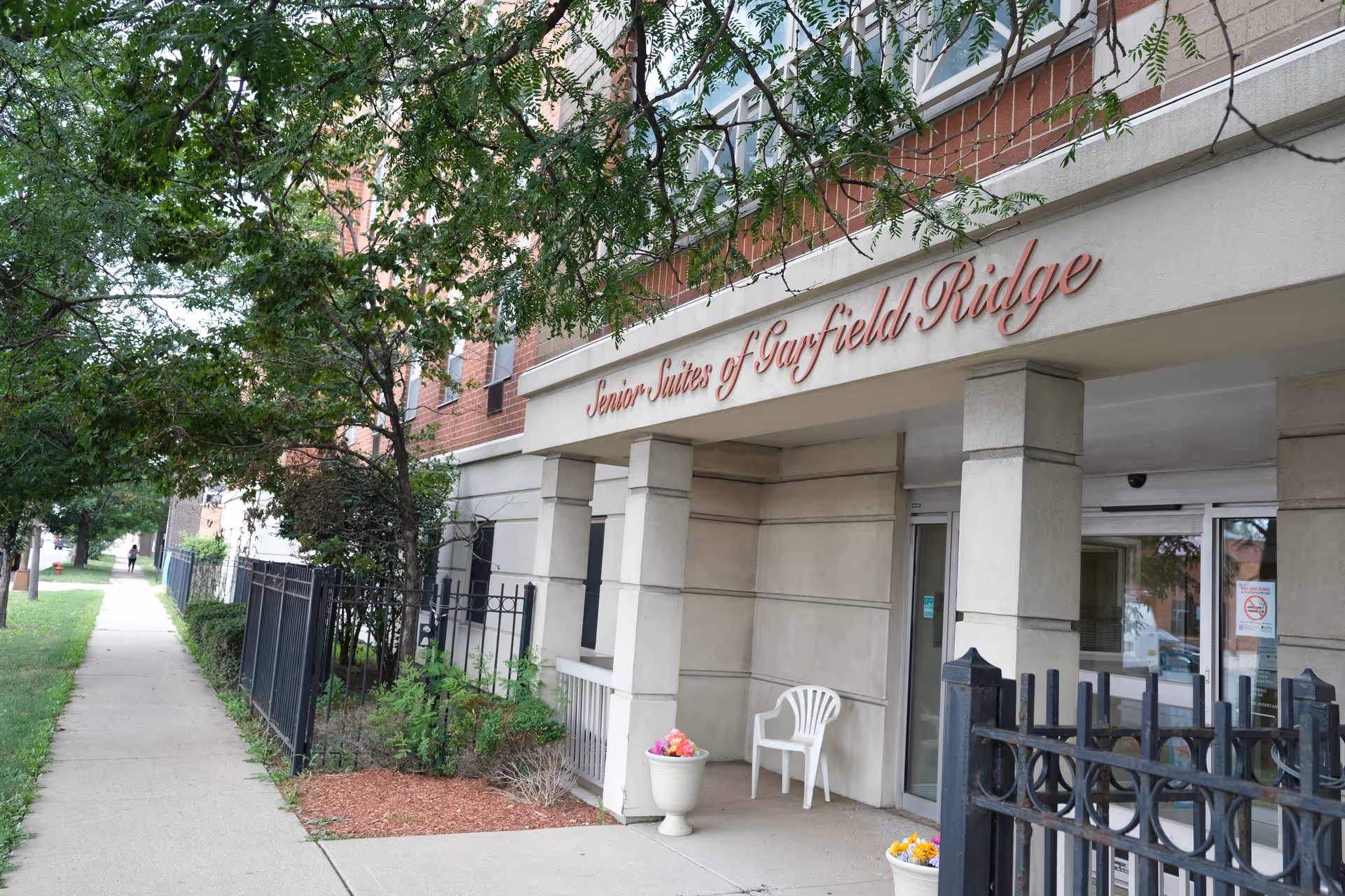 Entrance to the Senior Suites of Garfield Ridge building showing the name above columns, a sidewalk, black fence, and potted flowers.