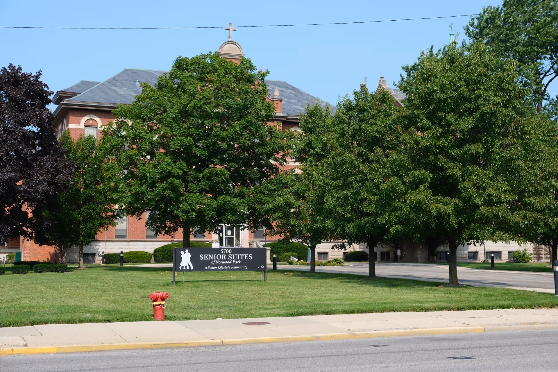 Front exterior of a brick Senior Suites of Norwood Park building with trees, lawn, and a sign.